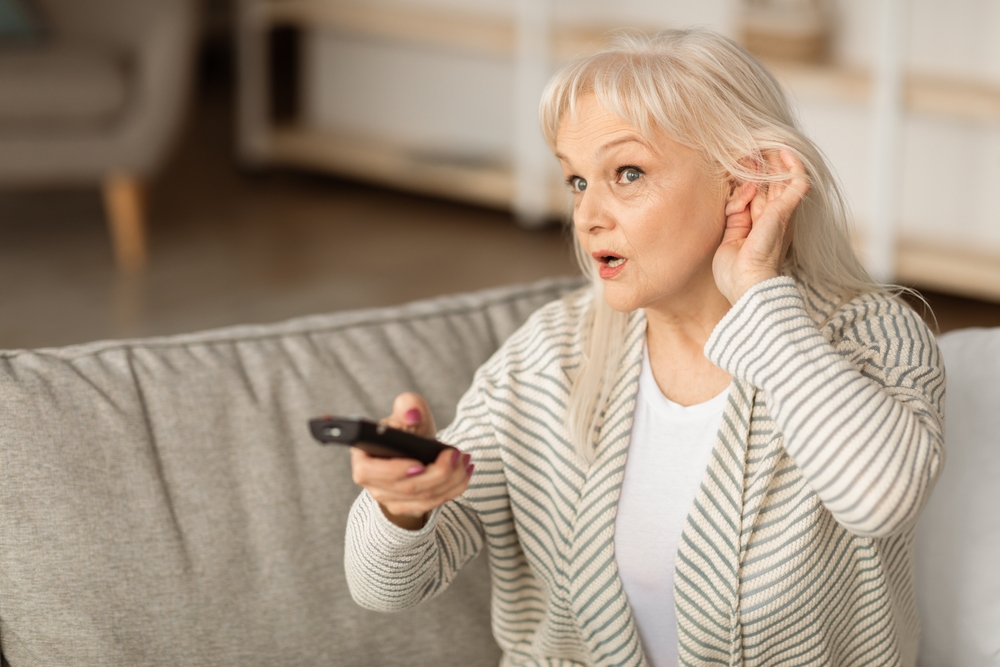 Older woman sitting on a couch, holding a TV remote and cupping her ear as if struggling to hear, with a concerned expression on her face.