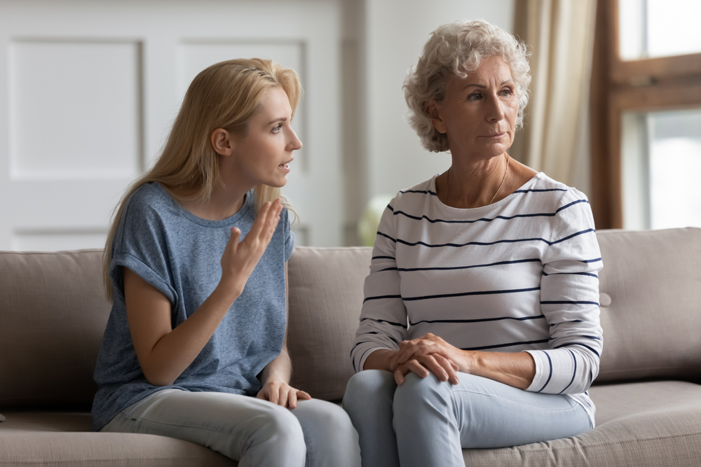 Young woman gesturing and speaking to an older woman who looks away with a serious expression as they sit on a couch, suggesting a tense or difficult conversation.