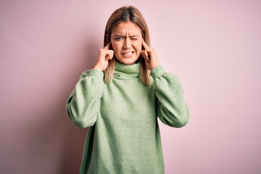 A woman in a green sweater stands against a pink background, closing her eyes and pressing her index fingers to her temples, appearing stressed or frustrated.