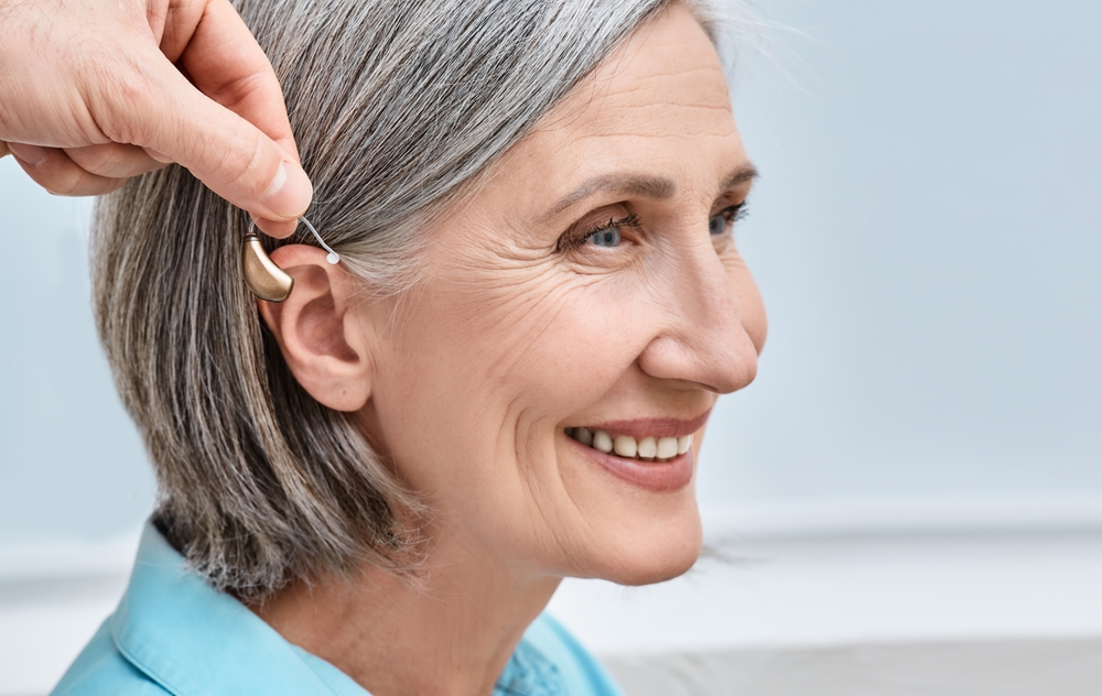 A smiling older woman with gray hair is having a hearing aid placed behind her ear by another person’s hand.