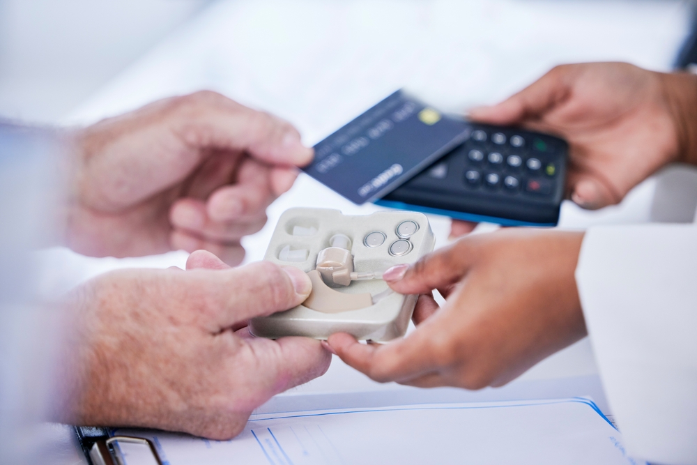 A person hands over a credit card to another person holding a payment terminal and a tray with hearing aid devices, suggesting a purchase or payment for medical equipment.