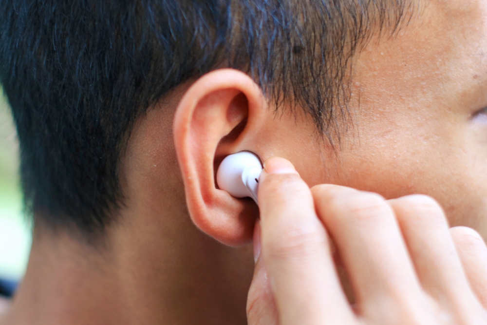 A person inserts a white wireless earbud into their ear, with their hand holding the earbud close to the ear. The close-up image focuses on the ear and earbud.